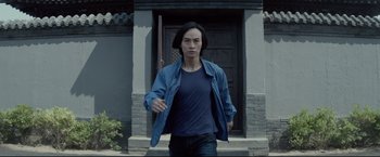 Movie still from “Man of Tai Chi” (2013), directed by Keanu Reeves – A man in a blue shirt holding a knife in front of a building; Medium shot, Low angle
