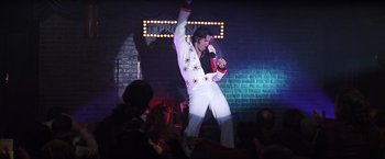 Movie still from “Man on the Moon” (1999), directed by Milos Forman – A man in white shirt and white pants holding a microphone on a stage; Wide shot, Low angle