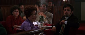 Movie still from “Man on the Moon” (1999), directed by Milos Forman – A woman sitting at a table with a man smoking a cigarette in the background; Medium shot, Over the shoulder angle