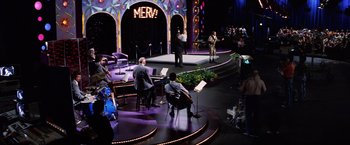 Movie still from “Man on the Moon” (1999), directed by Milos Forman – A group of people sitting on a stage playing instruments; Extreme Wide shot, High angle