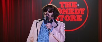 Movie still from “Man on the Moon” (1999), directed by Milos Forman – A man in a pink jacket holding a microphone and wearing sunglasses; Medium shot, Over the shoulder angle