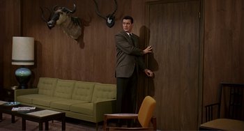 Movie still from “Man's Favorite Sport?” (1964), directed by Howard Hawks – A man in a suit leaning against a wall in a living room; Wide shot, Over the shoulder angle
