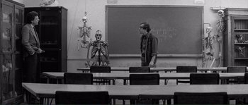 Movie still from “Manhattan” (1979), directed by Woody Allen – A man standing in front of a skeleton in front of a chalkboard in a class room; Wide shot, Over the shoulder angle