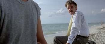 Movie still from “Manhunter” (1986), directed by Michael Mann – A man sitting on the beach with another man standing next to him; Medium shot, Over the shoulder angle