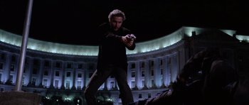 Movie still from “Manhunter” (1986), directed by Michael Mann – A man holding a gun in front of a large building at night; Wide shot, Low angle