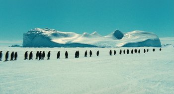 Movie still from “March of the Penguins” (2005), directed by Luc Jacquet – A group of penguins walking across a snow covered field; Extreme Wide shot, High angle