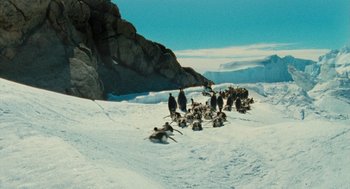Movie still from “March of the Penguins” (2005), directed by Luc Jacquet – A group of penguins walking across a snow covered ground; Extreme Wide shot, High angle