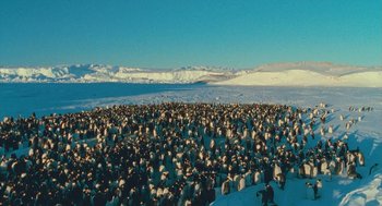 Movie still from “March of the Penguins” (2005), directed by Luc Jacquet – A large group of penguins gathered together in the snow; Extreme Wide shot, High angle