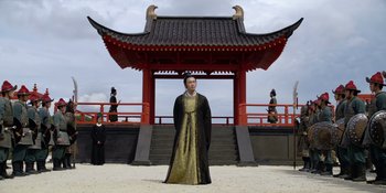 Movie still from “Marco Polo” (2014), created by John Fusco – A man dressed in oriental clothing standing in front of a building; Wide shot, Low angle
