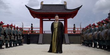 Movie still from “Marco Polo” (2014), created by John Fusco – A man dressed in traditional asian clothing standing in front of a pavilion; Wide shot, Low angle