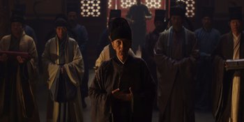 Movie still from “Marco Polo” (2014), created by John Fusco – An old man wearing a black hat in front of a group of people; Medium shot, Low angle