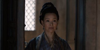 Movie still from “Marco Polo” (2014), created by John Fusco – A woman wearing a black hat and a blue and black dress; Close Up shot, Over the shoulder angle