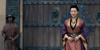 Movie still from “Marco Polo” (2014), created by John Fusco – A woman dressed in traditional asian clothing standing in front of a wooden door; Medium shot, Over the shoulder angle