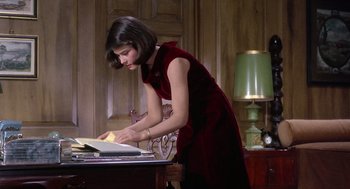 Movie still from “Marnie” (1964), directed by Alfred Hitchcock – A woman in a red dress looking at a book on top of a desk; Medium shot, High angle
