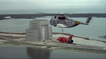 Movie still from “Marooned” (1969), directed by John Sturges – A helicopter is flying over a building near a body of water; Extreme Wide shot, High angle