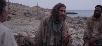 Movie still from “Mary Magdalene” (2018), directed by Garth Davis – A man with long hair and beard sitting on a rocky beach; Medium shot, Over the shoulder angle