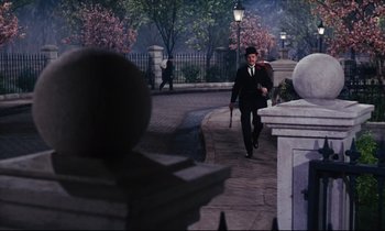Movie still from “Mary Poppins” (1964), directed by Robert Stevenson – A man walking down a street holding a cane; Wide shot, Over the shoulder angle
