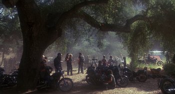 Movie still from “Mask” (1985), directed by Peter Bogdanovich – A group of people standing next to motorcycles under a tree; Wide shot, High angle