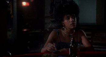 Movie still from “Mask” (1985), directed by Peter Bogdanovich – A woman sitting at a table in a dark room; Close Up shot, Over the shoulder angle
