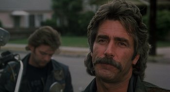 Movie still from “Mask” (1985), directed by Peter Bogdanovich – A man with long hair and a mustache is standing next to another man with long hair; Close Up shot, Over the shoulder angle