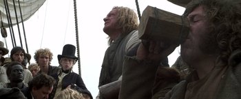 Movie still from “Master and Commander: The Far Side of the World” (2003), directed by Peter Weir – A man with long blonde hair is holding a wooden block; Medium shot, Low angle