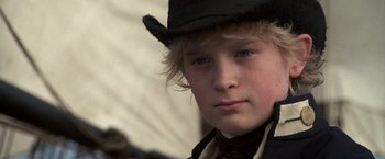 Movie still from “Master and Commander: The Far Side of the World” (2003), directed by Peter Weir – A young boy wearing a black hat and looking at the camera; Close Up shot, Over the shoulder angle