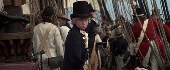 Movie still from “Master and Commander: The Far Side of the World” (2003), directed by Peter Weir – A young boy in a top hat standing in front of a group of people; Medium shot, Over the shoulder angle