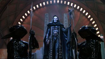 Movie still from “Masters of the Universe” (1987), directed by Gary Goddard – A statue of a man in a black outfit holding a spear; Wide shot, Low angle