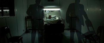 Movie still from “Matchstick Men” (2003), directed by Ridley Scott – A man laying in a hospital bed in a dark room; Wide shot, Low angle