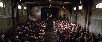 Movie still from “Matilda” (1996), directed by Danny DeVito – An audience is sitting in front of a stage with a man on the stage; Extreme Wide shot, High angle