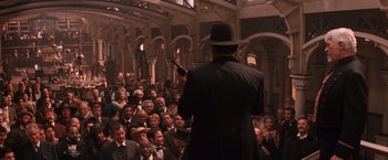 Movie still from “Maverick” (1994), directed by Richard Donner – A man in a black coat and a black hat is speaking to a group of people; Wide shot, High angle