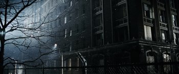 Movie still from “Max Payne” (2008), directed by John Moore – A person standing on a fence in front of a tall building; Extreme Wide shot, Low angle