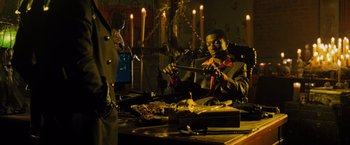 Movie still from “Max Payne” (2008), directed by John Moore – A man holding a knife in his hand while sitting at a table; Medium shot, Low angle