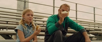 Movie still from “McFarland, USA” (2015), directed by Niki Caro – A man and a little girl sitting on bleachers eating hot dogs; Medium shot, Over the shoulder angle