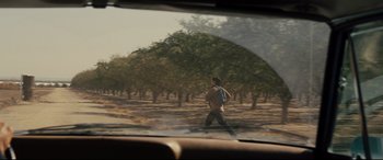 Movie still from “McFarland, USA” (2015), directed by Niki Caro – A man walking across a dirt road near a grove of trees; Wide shot, Over the shoulder angle