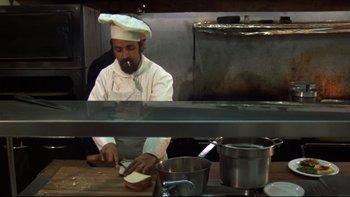 Movie still from “Mean Streets” (1973), directed by Martin Scorsese – A man wearing a chef's hat cutting a piece of bread; Medium shot, Low angle