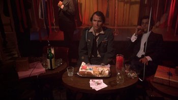 Movie still from “Mean Streets” (1973), directed by Martin Scorsese – A man sitting in front of a cake on top of a table; Medium shot, Over the shoulder angle