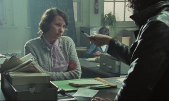 Movie still from “Meantime” (1983), directed by Mike Leigh – A woman sitting at a table in front of a man holding a cell phone; Medium shot, Over the shoulder angle