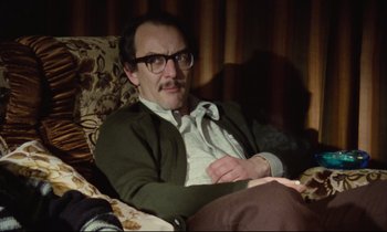 Movie still from “Meantime” (1983), directed by Mike Leigh – A man sitting on a couch with his arms crossed; Medium shot, High angle