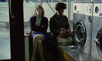 Movie still from “Meantime” (1983), directed by Mike Leigh – Two women sitting on a bench next to a washing machine; Medium shot, Low angle