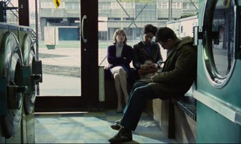 Movie still from “Meantime” (1983), directed by Mike Leigh – A man sitting on a bench next to two other people; Wide shot, High angle