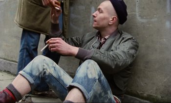 Movie still from “Meantime” (1983), directed by Mike Leigh – A man sitting on the ground smoking a cigarette; Medium shot, Overhead angle