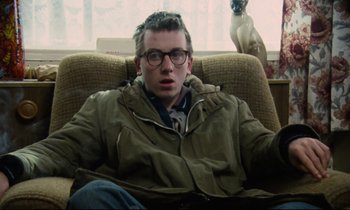 Movie still from “Meantime” (1983), directed by Mike Leigh – A man sitting in a chair with a cat in the background; Medium shot, Low angle