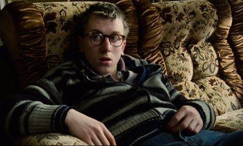 Movie still from “Meantime” (1983), directed by Mike Leigh – A man sitting on top of a couch wearing glasses; Medium shot, High angle