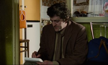 Movie still from “Meantime” (1983), directed by Mike Leigh – A man in a brown jacket is writing on a tablet; Medium shot, Over the shoulder angle