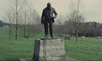 Movie still from “Meantime” (1983), directed by Mike Leigh – A statue of a man standing on top of a rock in a field; Wide shot, Low angle