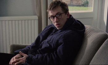 Movie still from “Meantime” (1983), directed by Mike Leigh – A man sitting on top of a couch wearing glasses; Medium shot, High angle