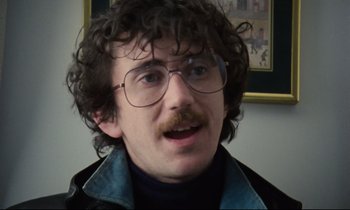 Movie still from “Meantime” (1983), directed by Mike Leigh – A man with curly hair wearing glasses and a black jacket; Close Up shot, Over the shoulder angle