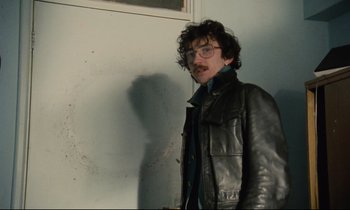 Movie still from “Meantime” (1983), directed by Mike Leigh – A man in a black leather jacket standing in front of a wall; Medium shot, Low angle