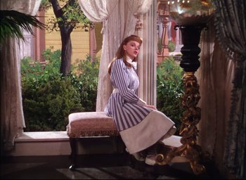 Movie still from “Meet Me in St. Louis” (1944), directed by Vincente Minnelli – A woman sitting on top of an ottoman in a living room; Wide shot, Low angle